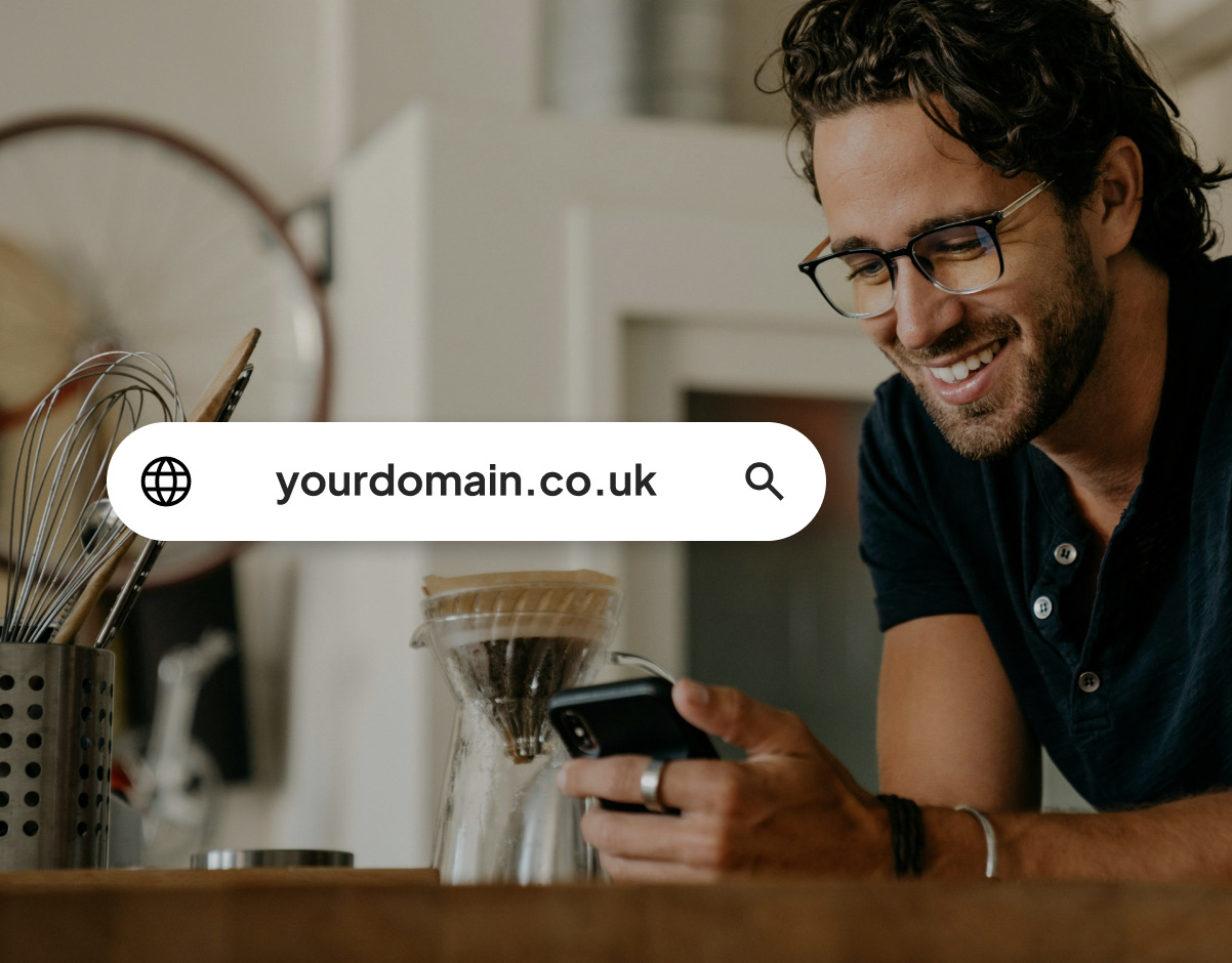 A man smiling at his phone in a kitchen, with a browser bar reading yourdomain.co.uk